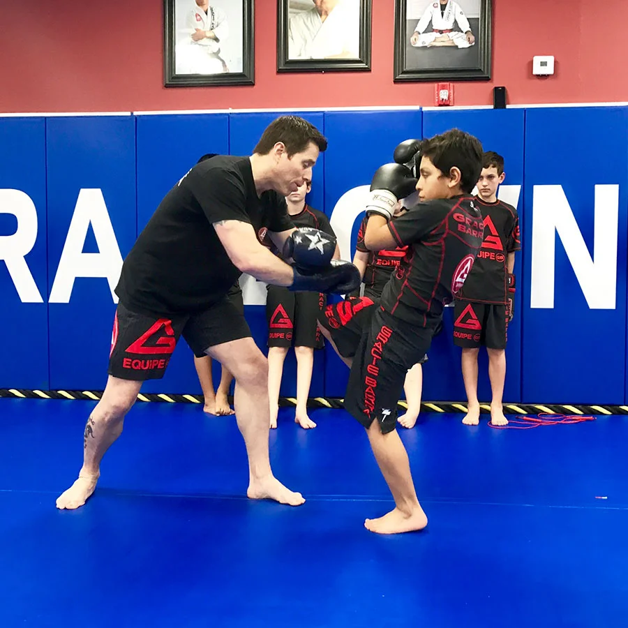 Instructor teaches a boy boxing in a gym with other children watching, all wearing matching attire, emphasizing self-defense techniques.