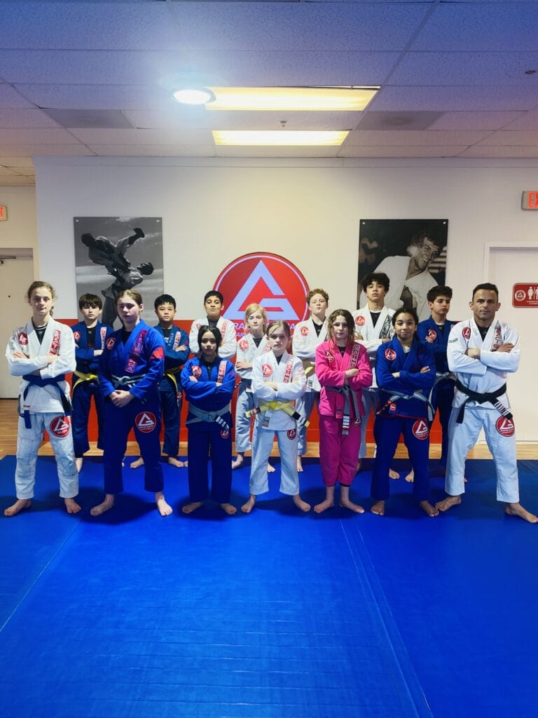 A group of people in martial arts uniforms stand on a blue mat, proudly posing in a dojo adorned with jiu jitsu posters on the walls, ready to master self-defense techniques.