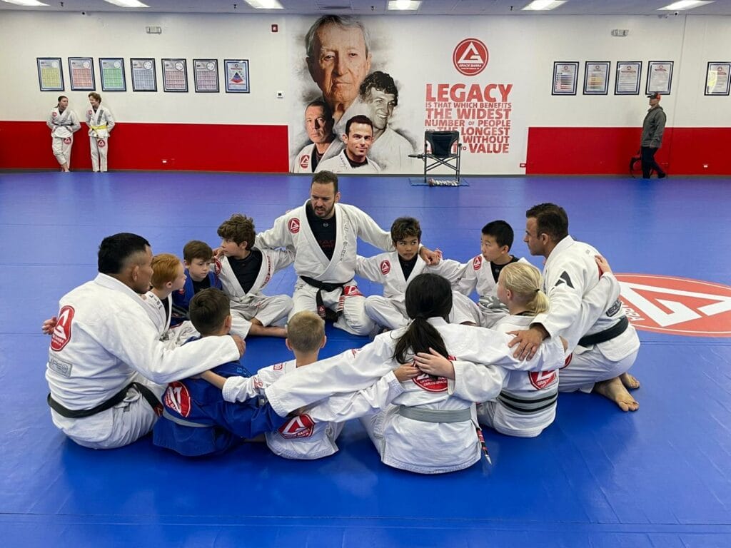 A group of jiu-jitsu students and their instructor sit in a circle on a blue mat, wearing white gis, in a martial arts training room with posters and images on the walls. Ready to take your skills to the next level? Join us today!