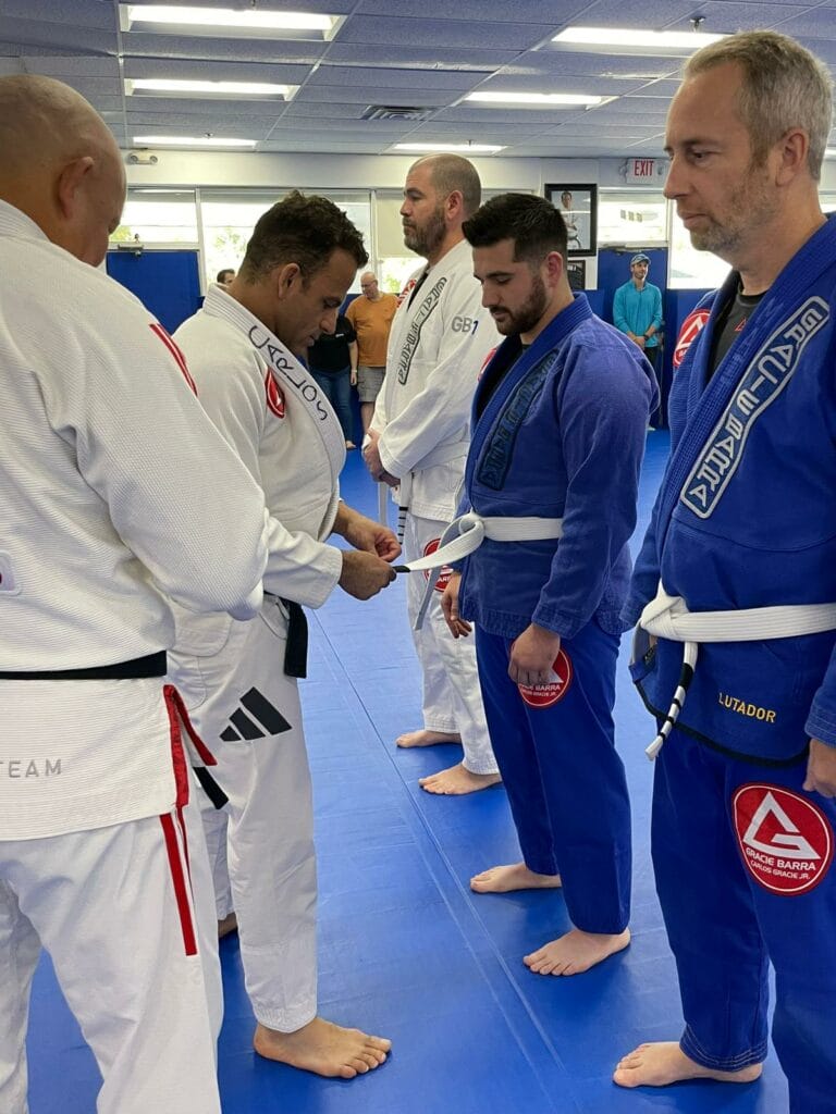 A group of people in martial arts uniforms stands in a line on a blue mat, participating in the GB1 Program. A man in a white gi is tying a belt around another person's waist.